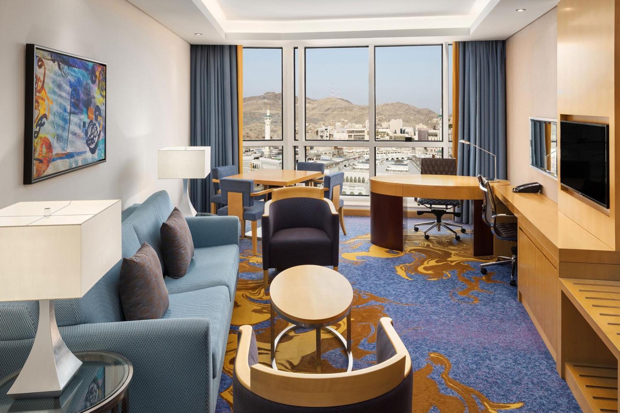 Sheraton Makkah Jabal Al Kaaba Hotel Mecca Exterior photo The photo shows a well-decorated hotel room featuring a modern and comfortable interior. There is a cozy seating area with a blue sofa and armchairs, complemented by a round coffee table. A desk with a chair is also present, ideal for work. The room 